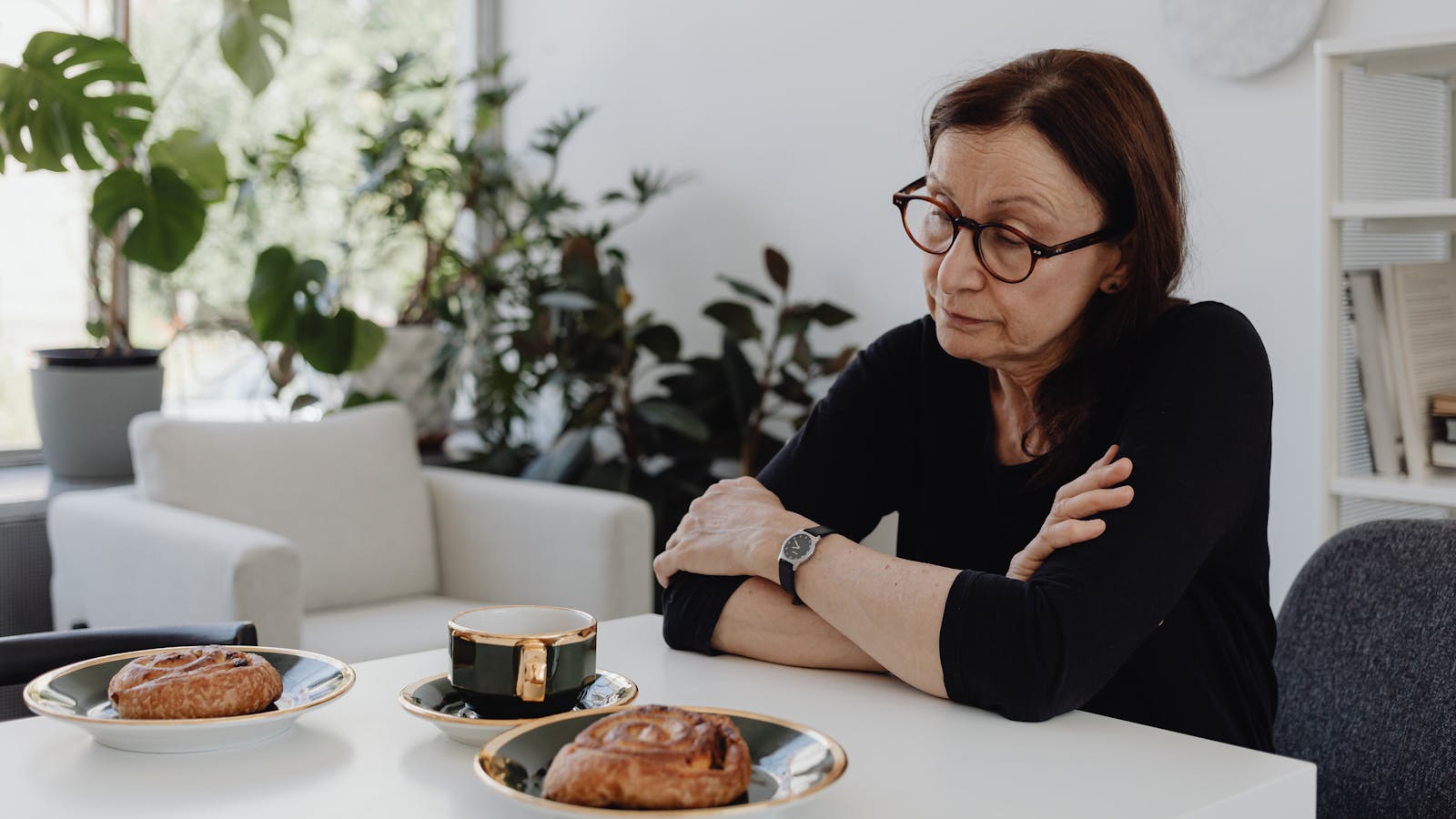 A woman with glasses sitting alone at a kitchen table, arms folded, looking down at a cup of tea